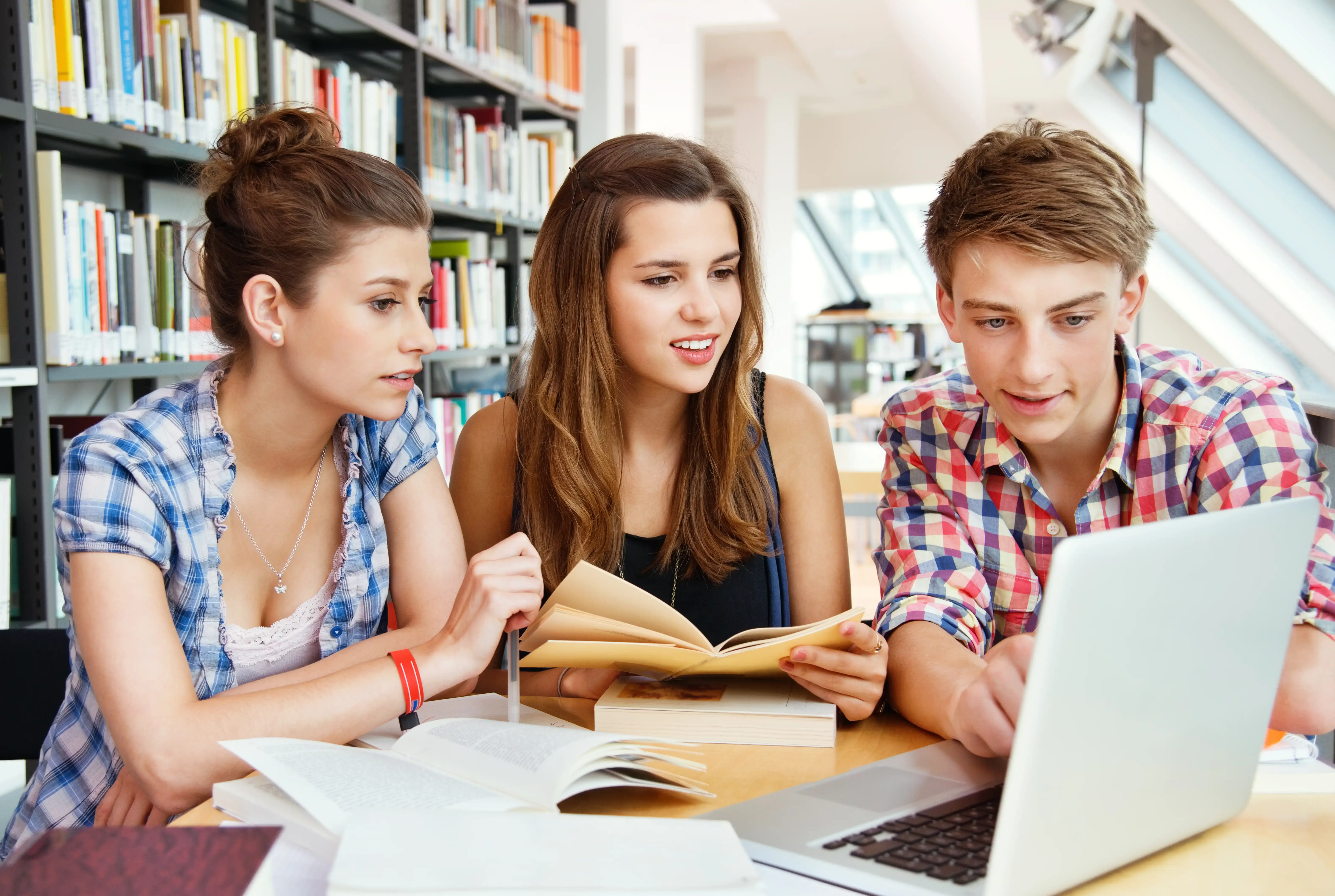 Students studying together at a library table, using books and a laptop for research and learning.